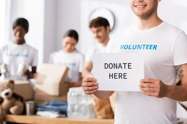 Selective focus of volunteer holding card with donate here lettering in charity center — Stock Photo