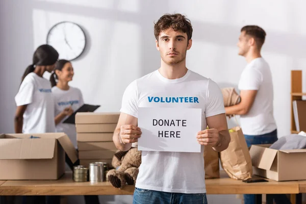 Selective focus of volunteer holding card with donate here lettering in charity center — Stock Photo