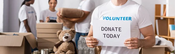 Horizontal image of volunteer holding card with donate today lettering near multiethnic people in charity center at background — Stock Photo