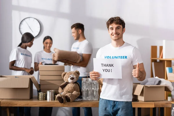 Focus selettivo di volontariato in possesso di carta con ringraziamento lettering e mostrando pollice in centro di beneficenza — Foto stock