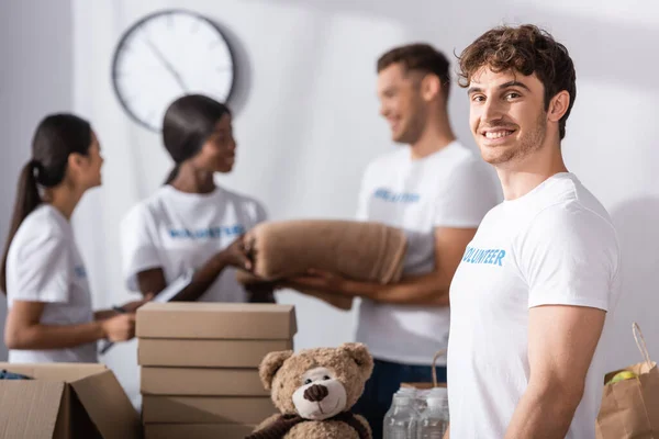 Selective focus of volunteer looking at camera near cardboard boxes and multiethnic people in charity center — Stock Photo