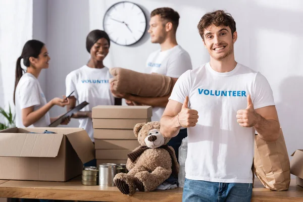 Enfoque selectivo del voluntario mostrando pulgares hacia arriba cerca de paquetes y personas multiétnicas en el centro de caridad - foto de stock