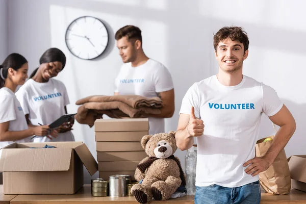 Selective focus of volunteer showing like near packages on table and multiethnic people in charity center — Stock Photo