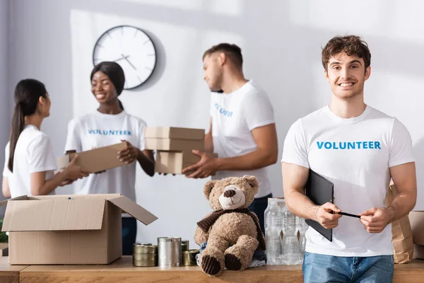 Selektiver Fokus von Freiwilligen, die Klemmbrett und Stift in der Nähe multiethnischer Menschen mit Paketen im Charity Center halten — Stockfoto