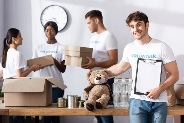 Selective focus of volunteer holding clipboard and soft toy near multiethnic people in charity center — Stock Photo