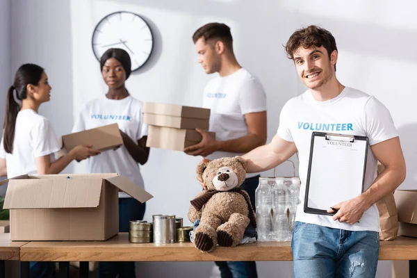 Concentration sélective du bénévolat tenant le presse-papiers et le jouet doux près des personnes multiethniques dans le centre de charité — Photo de stock