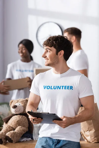 Selective focus of volunteer holding clipboard and pen near soft toy and paper bag in charity center — Stock Photo