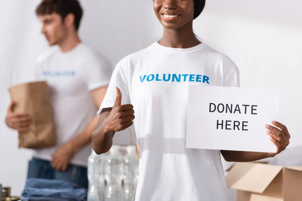 Selective focus of african american volunteer holding card with donate here lettering and showing like gesture — Stock Photo