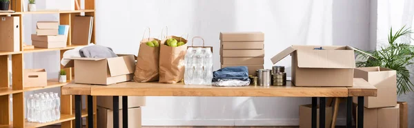 Panoramic shot of cardboard boxes, tin cans and clothes on table in charity center — Stock Photo