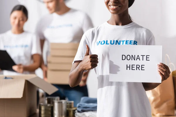 Foco seletivo do voluntário afro-americano mostrando como e segurando cartão com doar aqui lettering — Fotografia de Stock