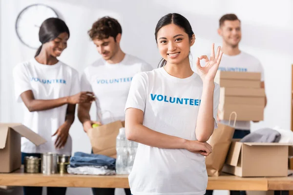 Enfoque selectivo de asiático voluntario mostrando ok gesto cerca de donaciones y personas multiétnicas en el centro de caridad en el fondo - foto de stock