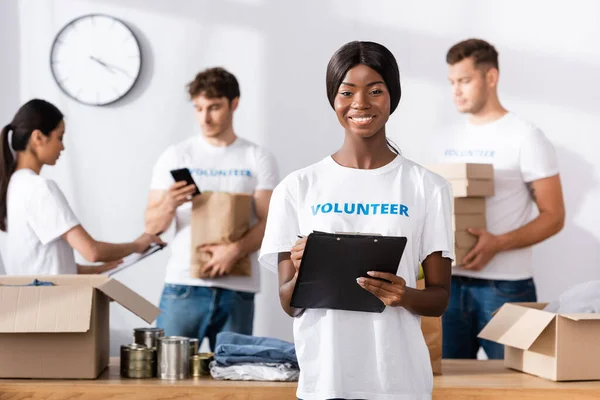 Selective focus of african american volunteer holding clipboard and looking at camera in charity center — Stock Photo