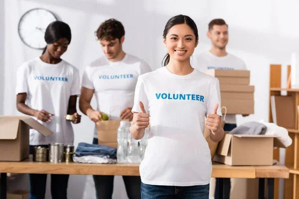 Enfoque selectivo de voluntarios asiáticos mostrando pulgares hacia arriba cerca de donaciones y personas multiétnicas en el centro de caridad - foto de stock