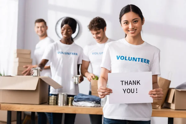 Selektiver Fokus einer asiatischen Frau, die in die Kamera blickt, während sie im Charity Center eine Karte mit Dankeschön-Schriftzug hält — Stockfoto