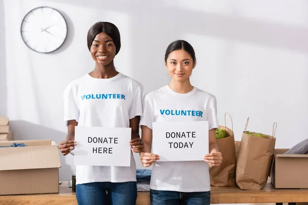 African american and asian volunteers holding cards with donate today and here lettering in charity center — Stock Photo