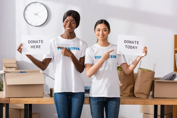 Voluntarios multiculturales señalando tarjetas con donaciones aquí y hoy en el centro de caridad - foto de stock