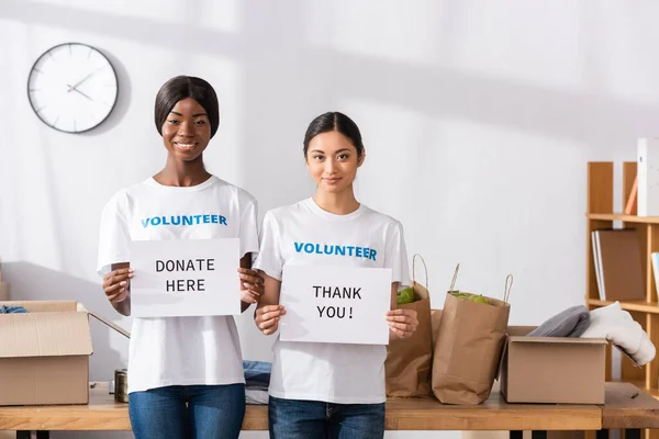 Voluntários multiétnicos segurando cartões com obrigado e doar aqui lettering no centro de caridade — Fotografia de Stock