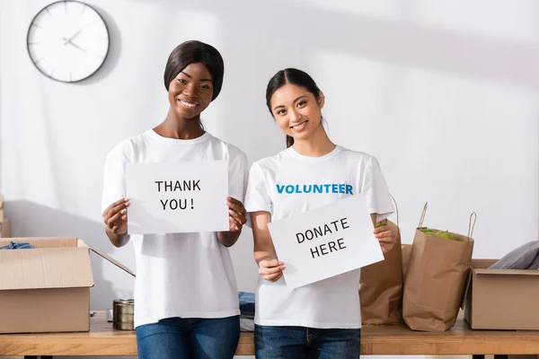 Multicultural volunteers holding cards with donate here and thank you lettering in charity center — Stock Photo