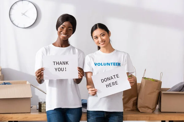 Voluntarios multiculturales sosteniendo tarjetas con donar aquí y gracias letras en el centro de caridad - foto de stock