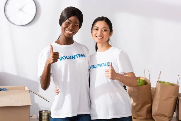 Multiethnic volunteers showing thumbs up near donations in charity center — Stock Photo