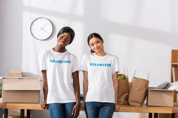 Multicultural volunteers smiling at camera near donations on table — Stock Photo