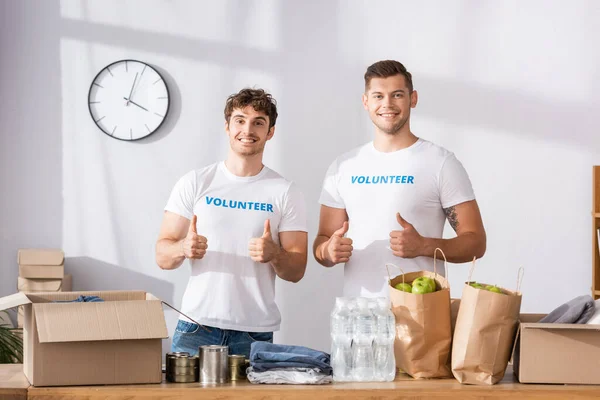 Voluntários mostrando polegares perto de pacotes e roupas na mesa no centro de caridade — Fotografia de Stock