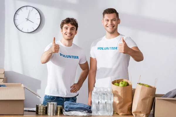 Jóvenes voluntarios mostrando pulgares cerca de paquetes, latas y ropa en la mesa - foto de stock