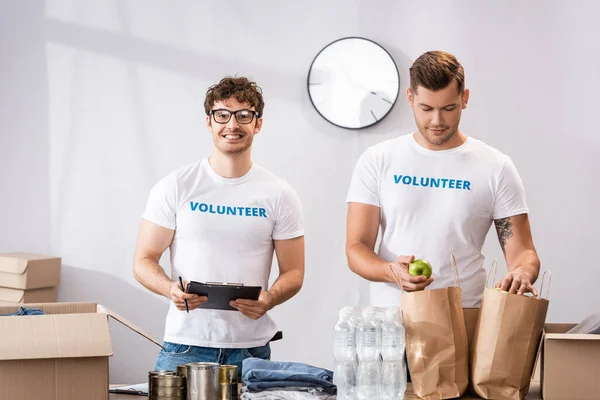 Selective focus of volunteers with clipboard and apple standing near donations on table — Stock Photo