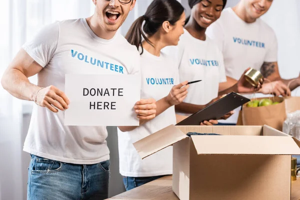 Selective focus of volunteer holding card with donate here lettering near donations and multicultural people — Stock Photo