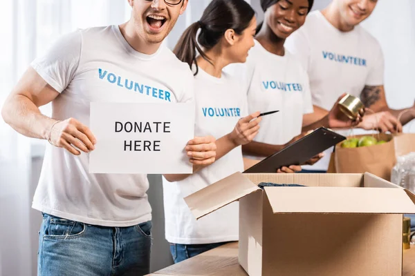 Foco seletivo do cartão de retenção voluntário com doar aqui lettering perto de doações e pessoas multiculturais — Fotografia de Stock