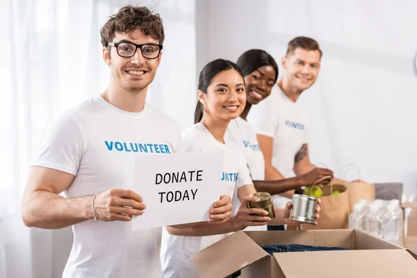 Selektiver Fokus multikultureller Freiwilliger, die in die Kamera blicken, während sie im Charity Center einen Ausweis mit Spendenaufdruck und Spenden in der Hand halten — Stockfoto