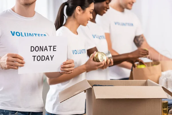 Selective focus of man holding card with donate today lettering near multiethnic volunteers and donations — Stock Photo