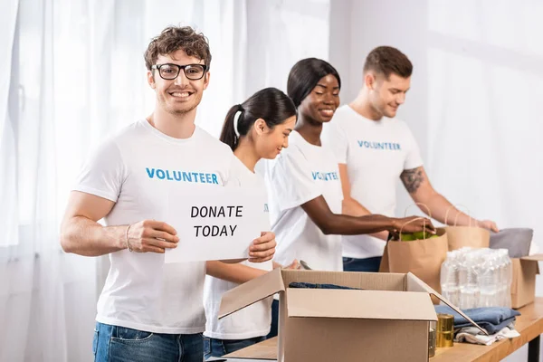 Selektiver Fokus des Mannes, der einen Ausweis mit Spendenaufdruck in der Nähe von Spenden und multikulturellen Freiwilligen in einem Wohltätigkeitszentrum hält — Stockfoto