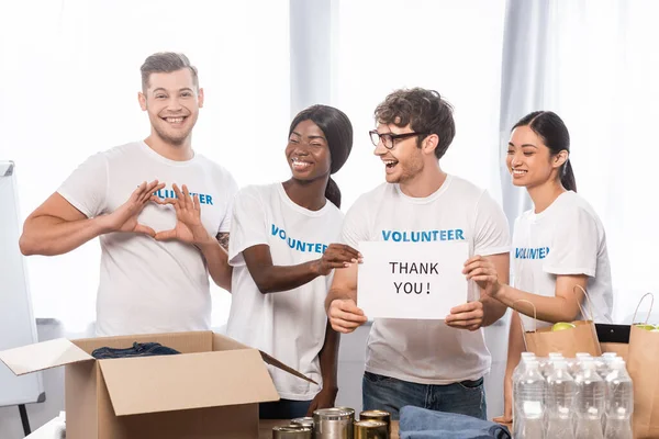 Selective focus of volunteer showing heart sign near laughing multiethnic people with lettering thank you on card and donations — Stock Photo