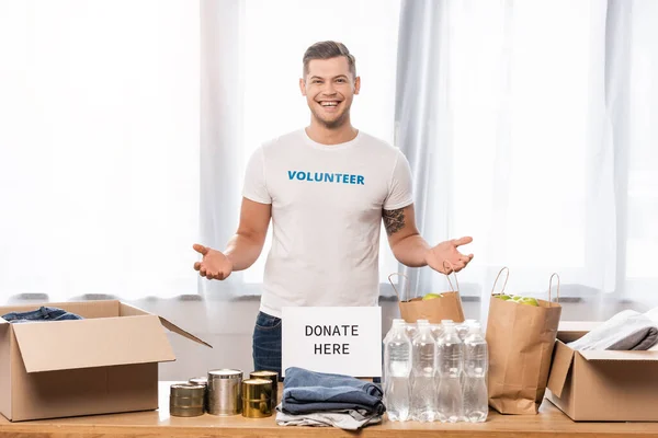 Volunteer pointing with hands near card with donate here lettering and donations on table — Stock Photo