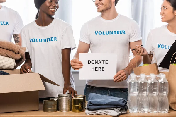 Мультиэтнические волонтеры держат карточку с пожертвованием здесь букв и пожертвований на столе — стоковое фото