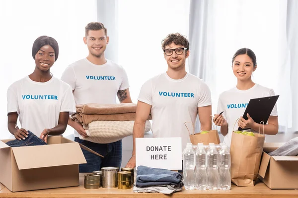 Multiethnic volunteers with clipboard and clothes standing near card with donate here lettering in charity center — Stock Photo