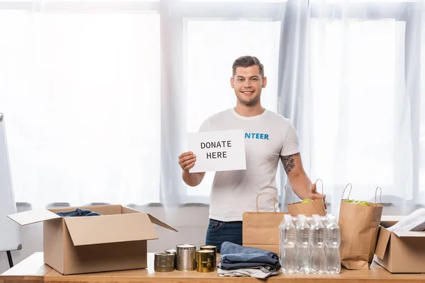 Jovem voluntário segurando cartão com doar aqui letras e doações na mesa — Fotografia de Stock