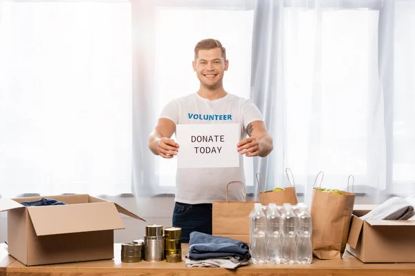 Volunteer holding card with donate today lettering near donations in charity center — Stock Photo