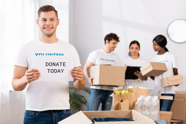 Selective focus of volunteer holding card with donate today lettering near packages and multiethnic people at background — Stock Photo