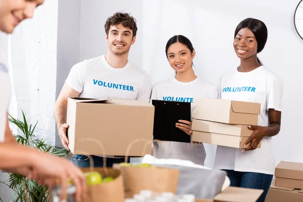 Focus selettivo di volontari multiculturali con scatole di cartone e appunti che guardano la fotocamera nel centro di beneficenza — Foto stock