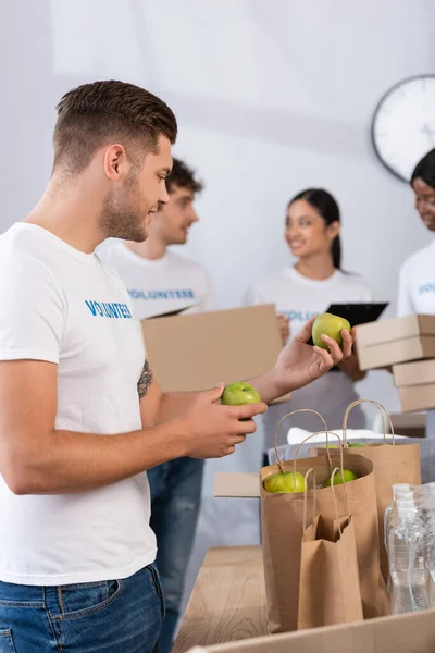 Foco seletivo do voluntário segurando maçãs perto de sacos de papel no centro de caridade — Fotografia de Stock
