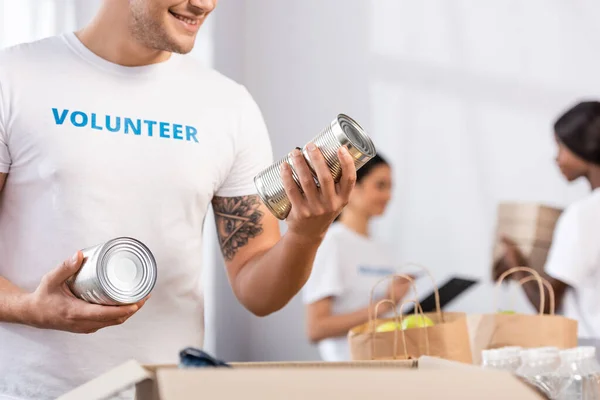 Foco seletivo do voluntário segurando latas de lata perto do pacote no centro de caridade — Fotografia de Stock