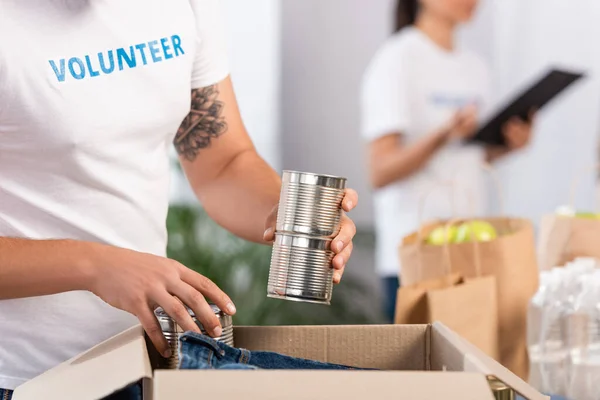 Vista recortada de voluntarios poniendo latas de lata en caja de cartón - foto de stock