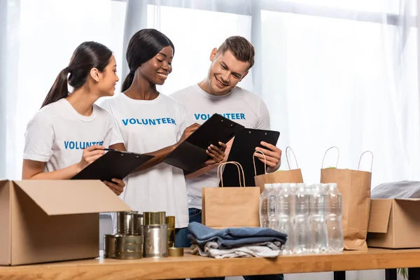 Foco seletivo de voluntários multiculturais olhando para pranchetas perto de doações e pacotes no centro de caridade — Fotografia de Stock