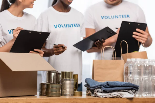 Vista cortada de voluntários multiétnicos com pranchetas perto de latas de lata, roupas e pacotes no centro de caridade — Fotografia de Stock