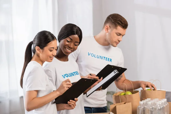 Selective focus of multicultural volunteers looking at clipboards near paper bags in charity center — Stock Photo