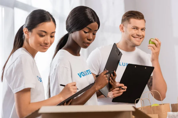 Selective focus of African american volunteer holding clipboard near people and packages in charity center — Stock Photo