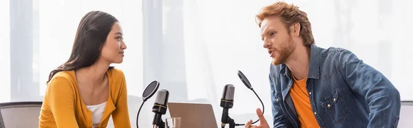 Молодая рыжая телеведущая разговаривает с брюнеткой азиаткой во время интервью, панорамный снимок — стоковое фото