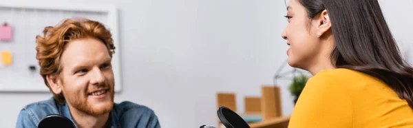 Selective focus of young redhead radio host interviewing brunette asian woman in studio, horizontal image — Stock Photo